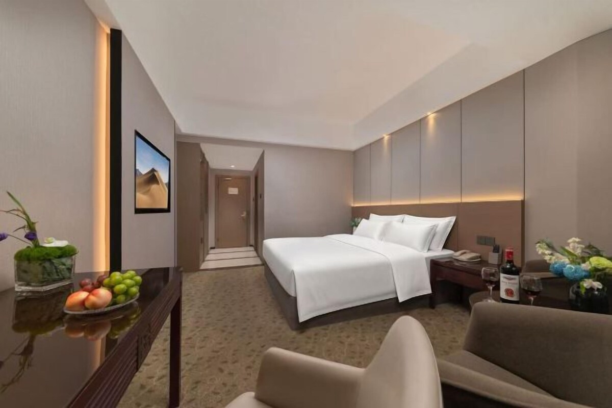 A hotel room with a bed, a desk with a fruit bowl, a chair, and a nightstand with a bottle.