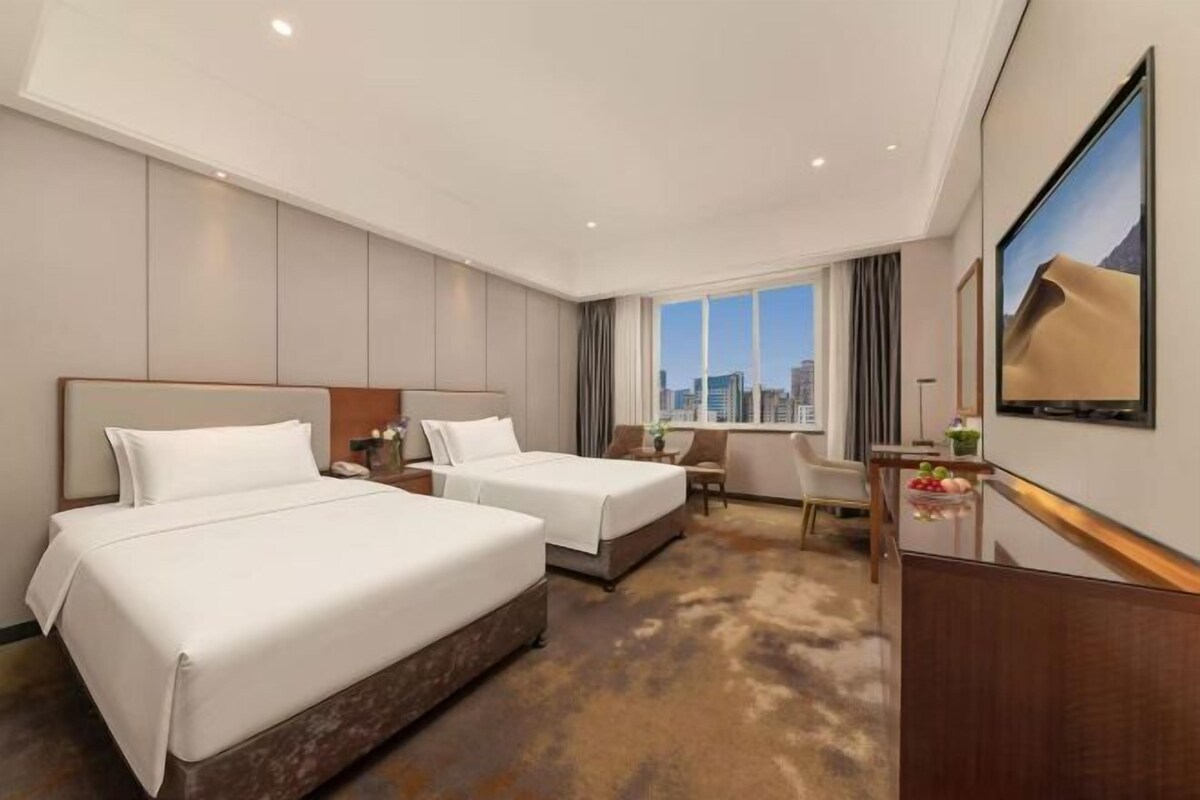 A hotel room with two beds, a TV, a desk, and a view of the city.
