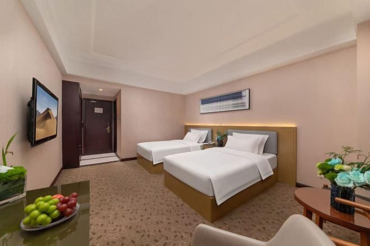 A hotel room with two beds, a desk with fruit, a TV, and a small table with flowers.