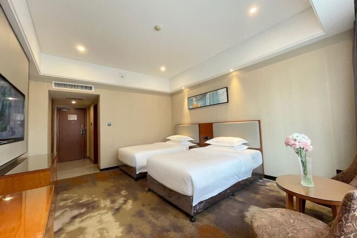 A hotel room with two beds, a small table with a vase, a sofa, a TV, and a door leading to another room.