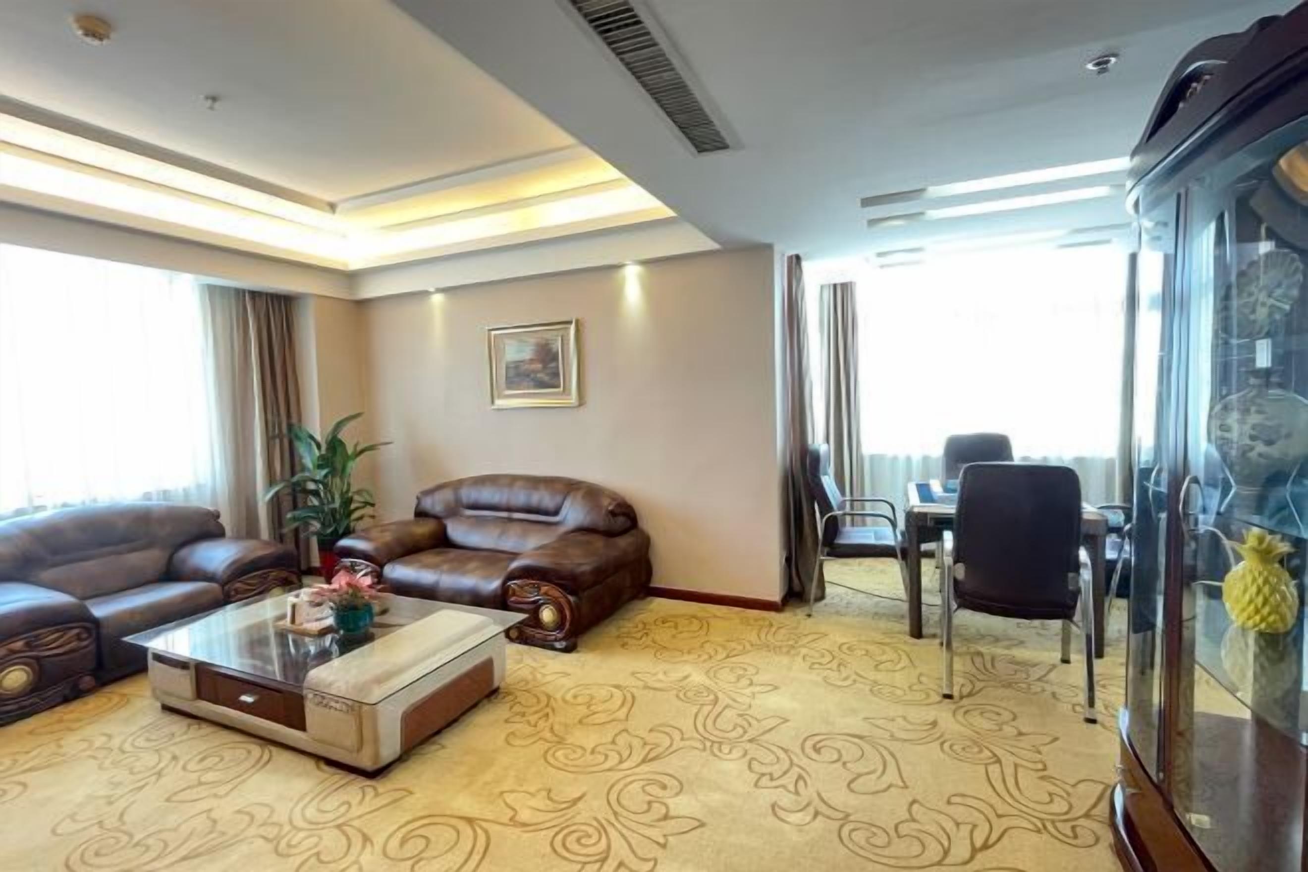 Habitación de hotel con sofá, una mesita de centro, una mesa de comedor con sillas, un ventanal amplio y un aparador de vidrio decorativo.