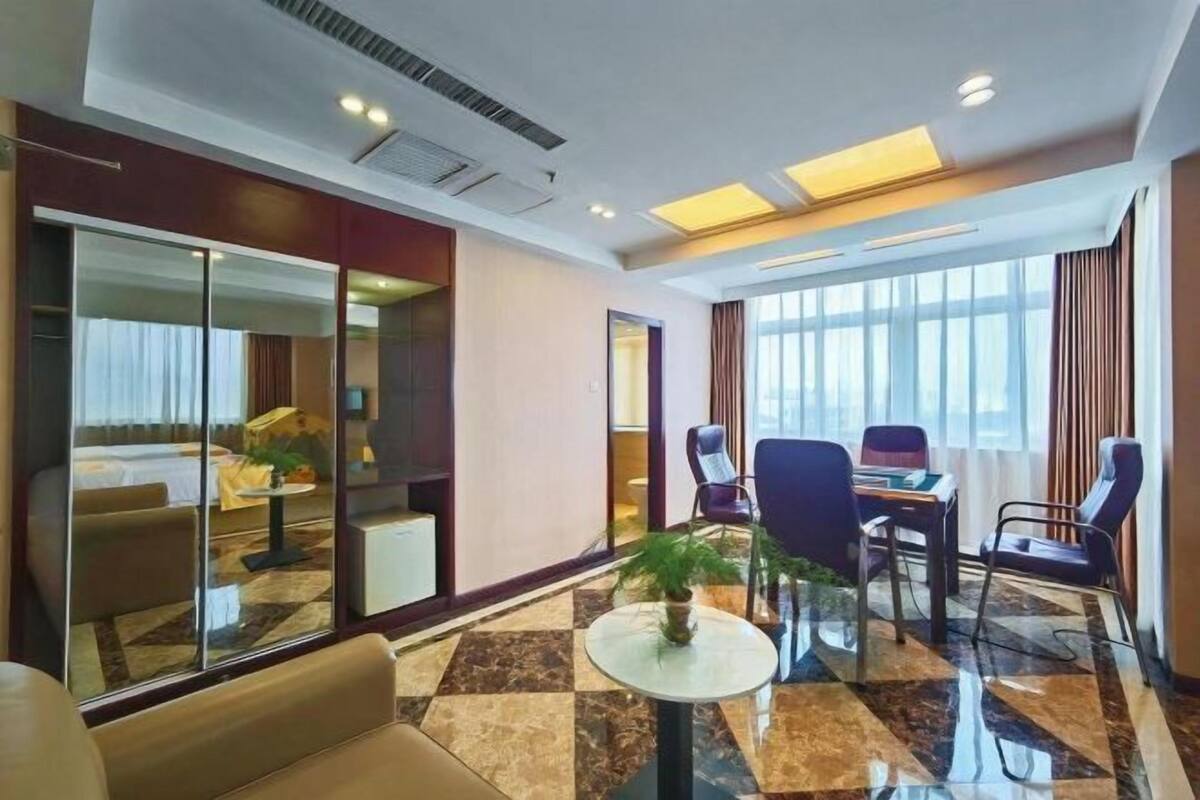 A hotel room with a bed, a seating area with chairs and a table, a large window with curtains, and a check-in counter.