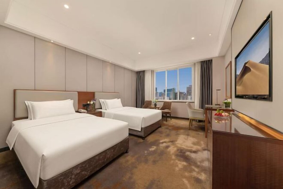 A hotel room with two beds, a desk, a chair, a TV, and a city view.