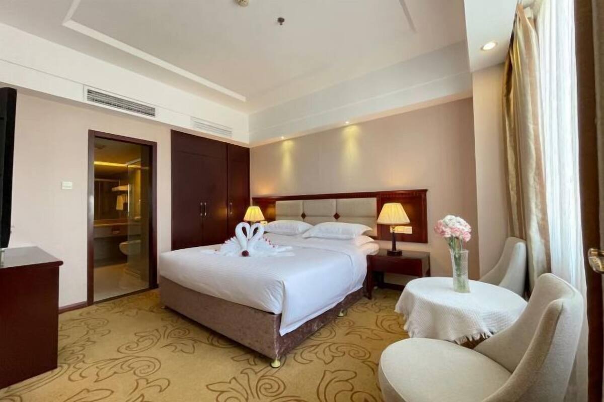 A hotel room with a large bed, bedside tables with lamps, a small round table with a vase, and a door leading to a bathroom.