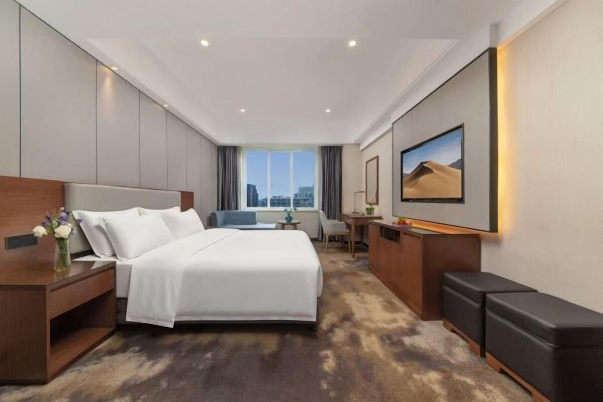 A hotel room with a large bed, a desk, a chair, and a view of the city through the window.