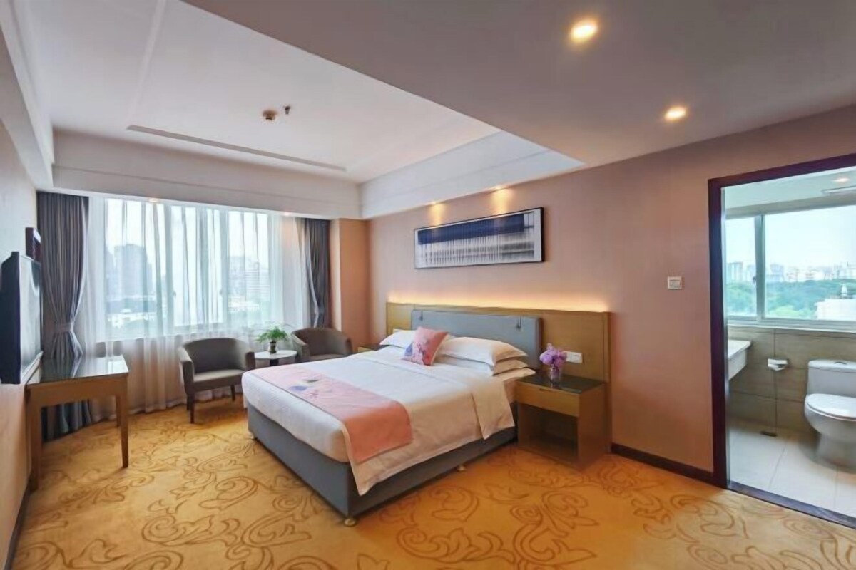 A hotel room with a bed, a desk, a chair, a nightstand, and a bathroom visible through a glass door.
