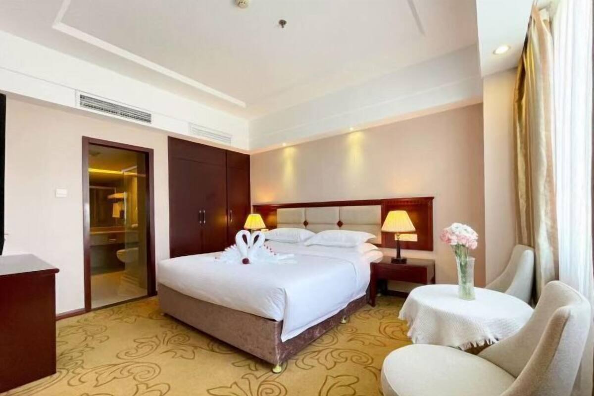A hotel room with a large bed, a desk, a chair, a nightstand, and a small table with a vase of flowers.
