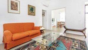 Una sala de estar con un sofá, una mesa de centro de vidrio y una alfombra estampada. Hay dos cuadros en la pared y una puerta que da a otra habitación.