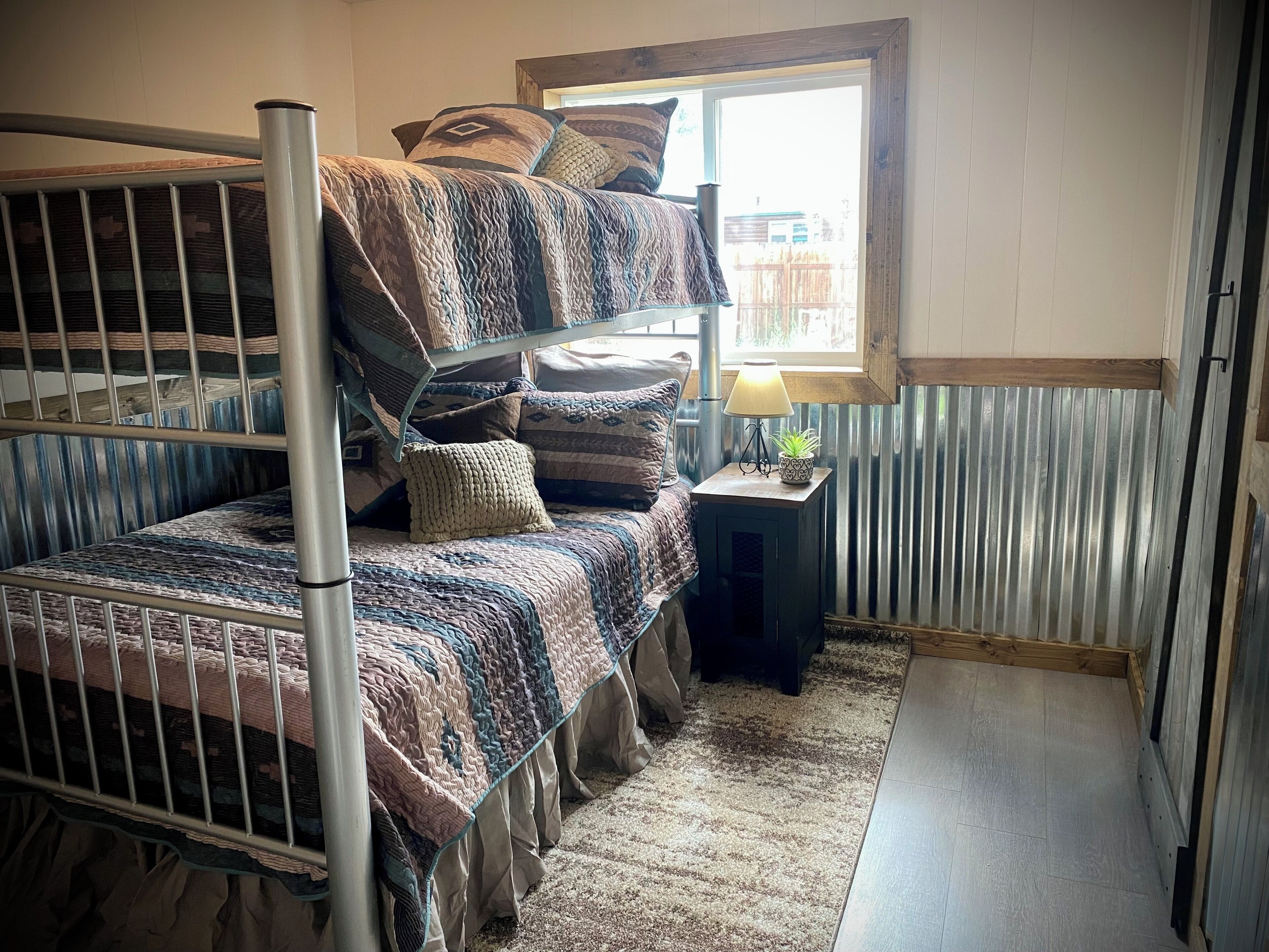 A bunk bed room with a window, a small table with a lamp, and a rug.