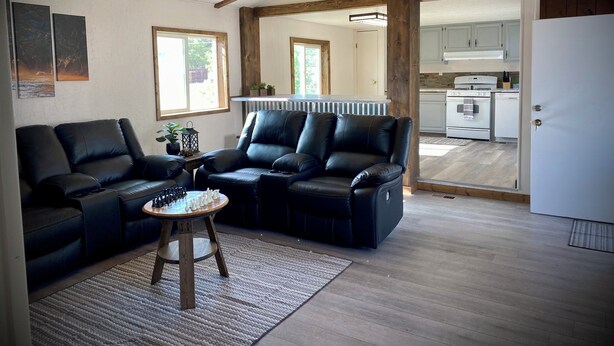A modern living room with a sectional sofa, a coffee table, and a fireplace. There are wooden beams on the ceiling, and a kitchen is visible in the background. - The Man Cave. Perfect for traveling workers and a great place for families  (Greybull)