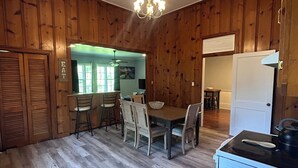 A wooden-paneled room with a dining table, chairs, a chandelier, and a kitchen area.