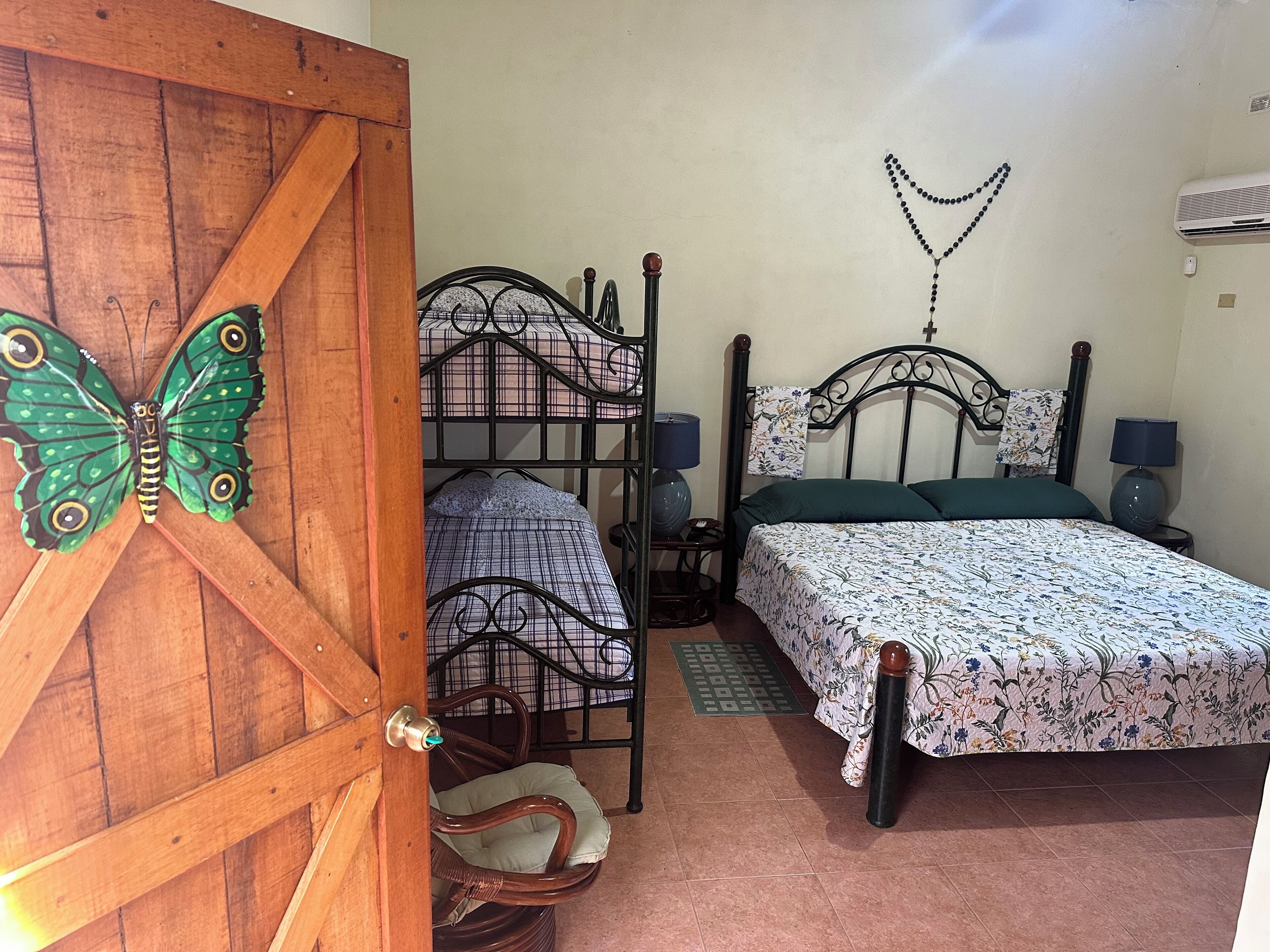 Une chambre avec une porte en bois, un lit recouvert d’une couvre-lit à motifs floraux, un deuxième lit recouvert d’une couvre-lit à carreaux, un climatiseur fixé au mur et une lampe fixée au mur.