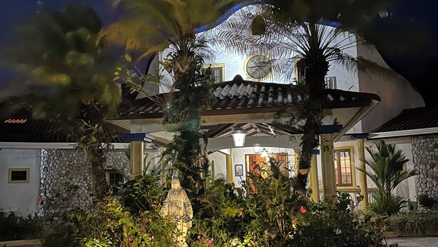 A well-lit building with a stone facade, a tiled roof, and a landscaped garden with a statue and palm trees. - Casa de Campo (El Valle de Antón)
