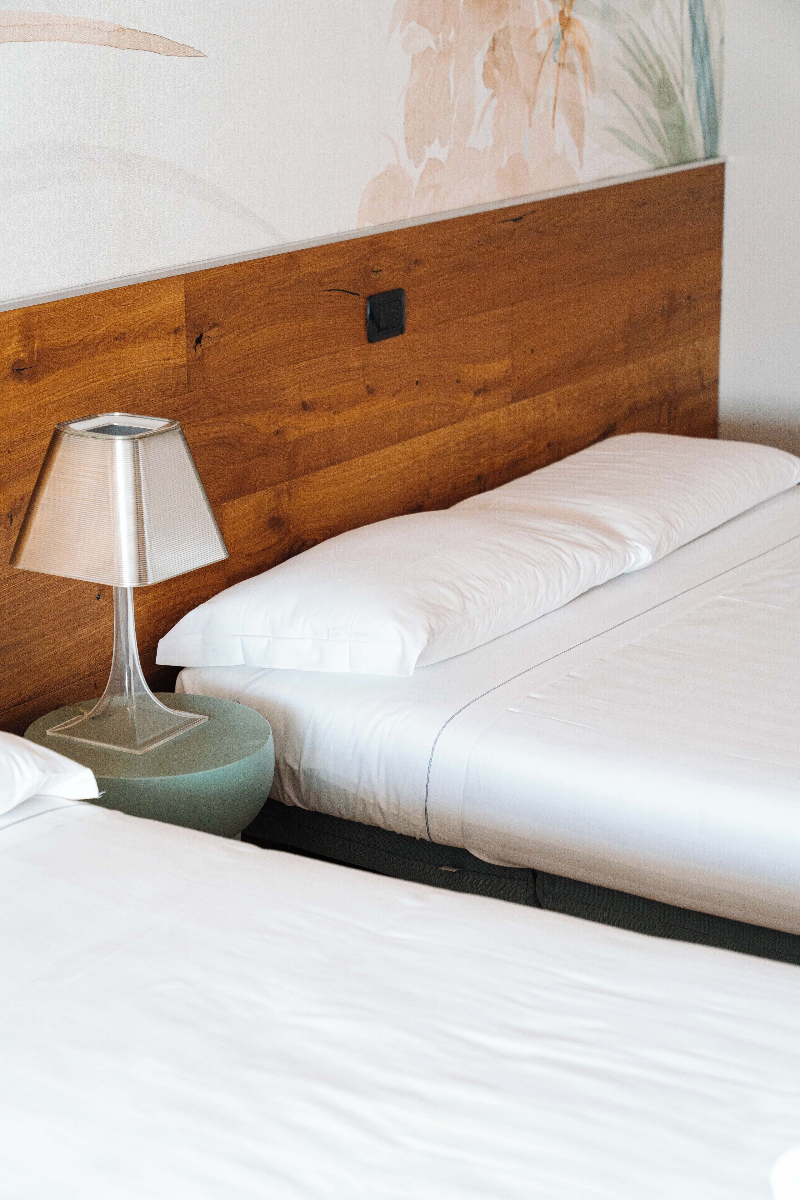 A bed with white linens, a wooden headboard, and a lamp on a round nightstand.