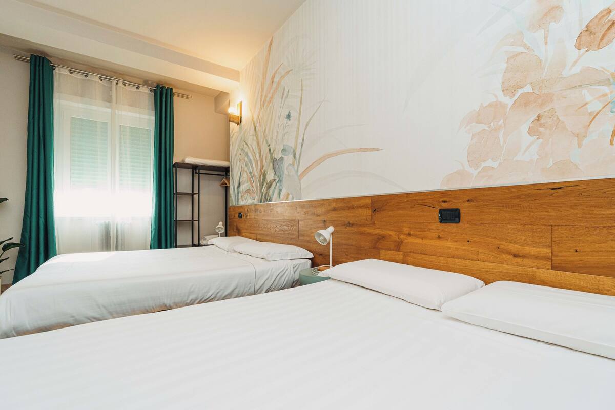 Hotellihuone, jossa on kaksi sänkyä, puinen pääty, seinämaalaus ja ikkuna, jossa on verhot.