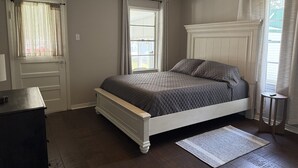 Ein Schlafzimmer mit Bett, Deckenventilator, Fenster mit Vorhängen, Tür und kleinem Tisch.