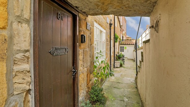 Un callejón estrecho con una puerta de madera, paredes de piedra y un techo de tejas, visible a través de un vano abierto.
