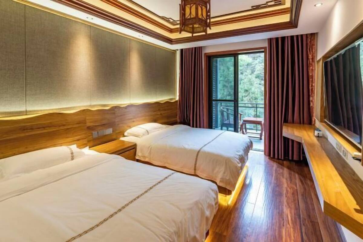 A hotel room with two beds, wooden headboards, a large window with curtains, and a balcony with a chair.