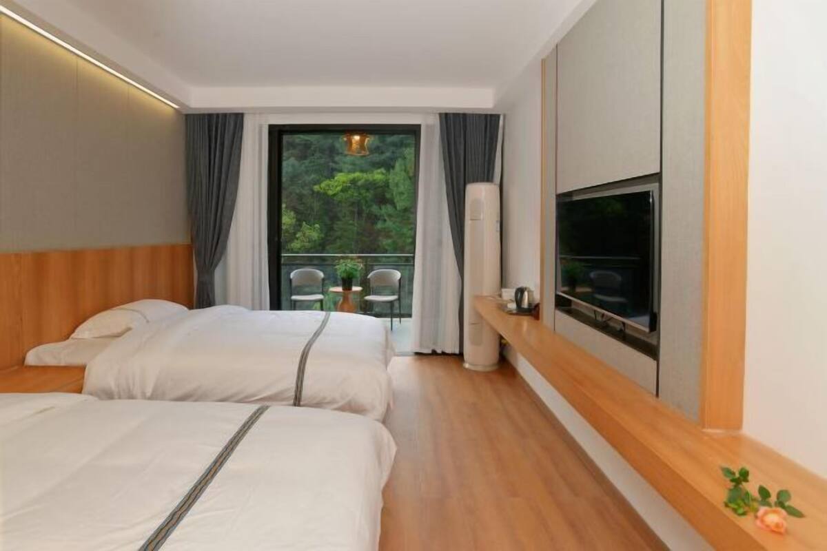 A hotel room with two beds, a TV, a balcony with chairs, and a wooden shelf.