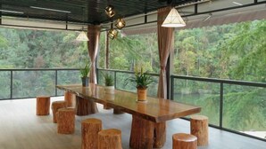 A spacious room with a large wooden table surrounded by stools, large windows offering a view of a forested area, and hanging pendant lights. - Shanlin Holiday Hotel (Chongqing)