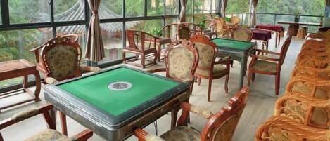 A room with a pool table, wooden chairs, and a view of greenery through large windows.