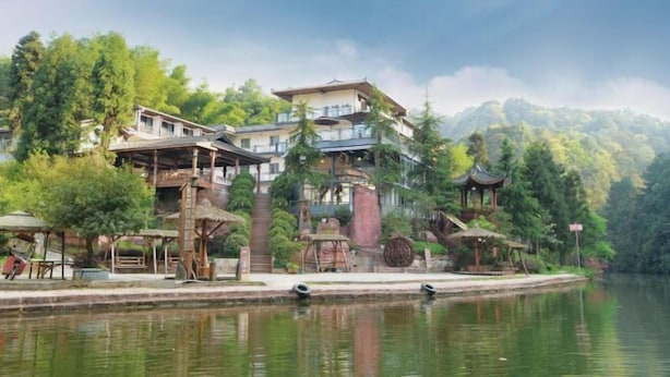 A traditional-style building with multiple levels, a water body in the foreground, and lush greenery in the background. - Shanlin Holiday Hotel (Chongqing)