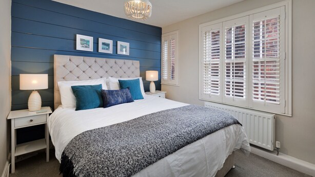 A bedroom with a large bed, two bedside tables, a chandelier, and a window with shutters. - The Gull's Nest (Whitby)