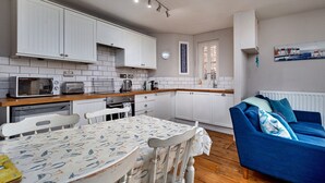 A compact kitchen with white cabinets, a microwave, a toaster, and a dining table with a printed tablecloth. - The Gull's Nest (Whitby)