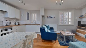 A modern kitchen with white cabinets, a dining table with a patterned tablecloth, and a blue sofa in a living area with a television and a window with blinds. - The Gull's Nest (Whitby)