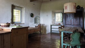 Ein rustikaler Raum mit einem hölzernen Bett, einem Holztisch und einem grünen Stuhl. Es gibt Fenster mit Gitterstäben, einen Holzbalken an der Decke und eine Hängelampe.
