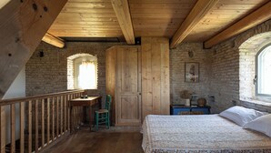Ein rustikales Schlafzimmer mit Holzbalkendecke, einem Bett mit gemustertem Überwurf, einem Holztisch, einem grünen Stuhl und einer Ziegelwand.