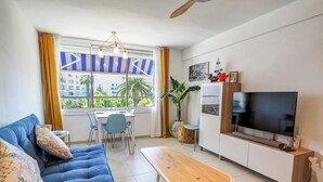 A modern living room with a blue sofa, a wooden coffee table, a dining area with chairs, a TV, and a ceiling fan. - Balcis Apartment by Hello Homes Sitges (Sitges)