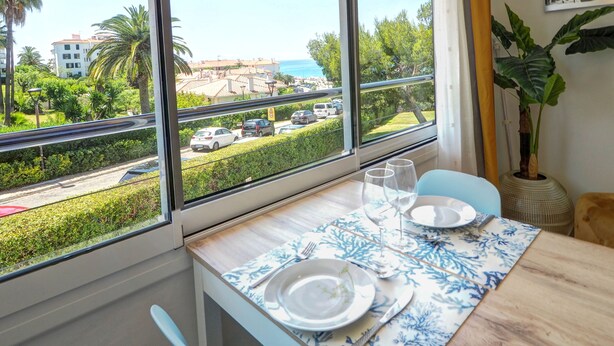 A dining area with a table set for two, a large window with a view of a street and buildings, and a potted plant on the windowsill. - Balcis Apartment by Hello Homes Sitges (Sitges)