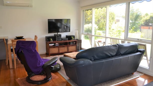A living room with a sofa, a television, a dining area with a table and chairs, and large windows with a view of greenery outside.
