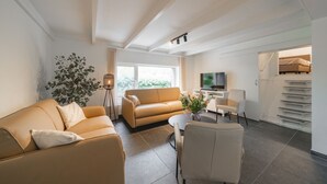 A modern living room with a sofa, a coffee table, and a TV. There is a plant and a window with curtains.
