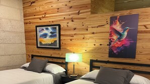 A room with two beds, wooden walls, a painting of a hummingbird, and a lamp.