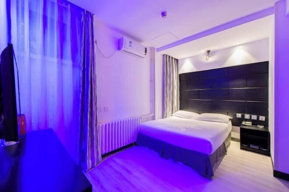 A hotel room with a bed, a bedside table, a chair, a radiator, and a wall-mounted air conditioning unit.