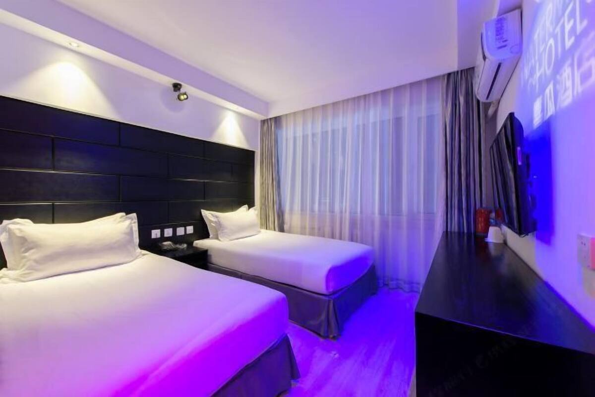 A hotel room with two beds, a flat-screen TV mounted on the wall, and a window with sheer curtains.