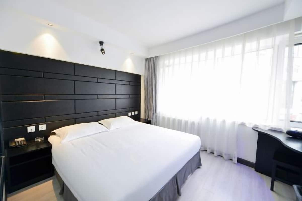A modern hotel room with a large bed, a desk, and a window with sheer curtains.