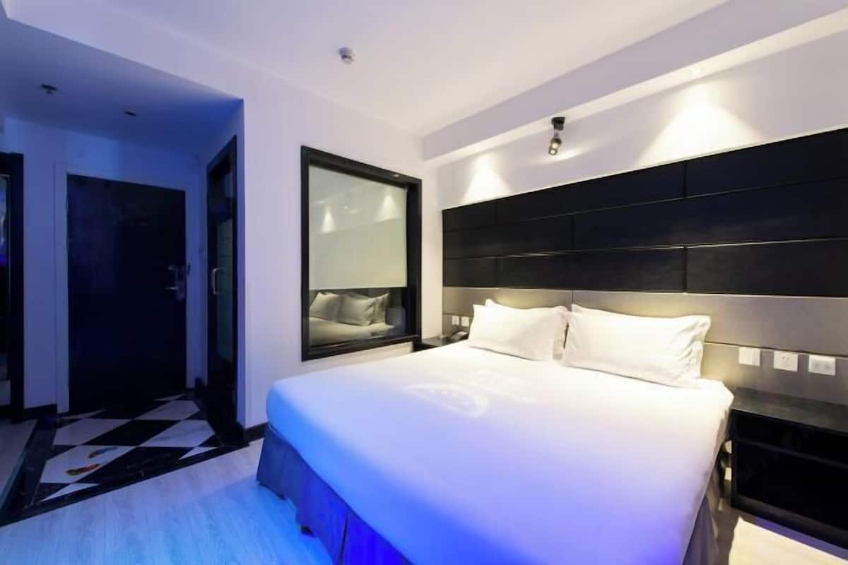 A modern hotel room with a large bed, a nightstand, a window with blinds, and a check-in desk.