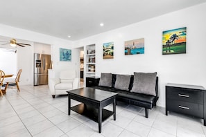 A modern living room with a sectional sofa, a coffee table, and a black side table. There are bookshelves with books and decorative items, and a ceiling fan. - Bungalow with large backyard, 5 min from beach (Pompano Beach)