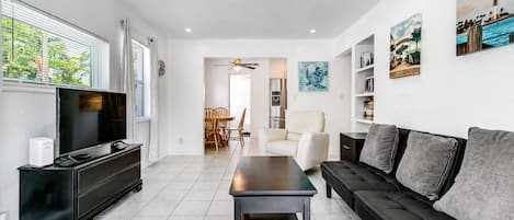 Un salon moderne avec un téléviseur à écran plat, un canapé foncé, une table basse et un coin repas à l’arrière-plan.