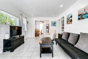 A modern living room with a black sofa, a coffee table, a TV, and a dining area in the background.