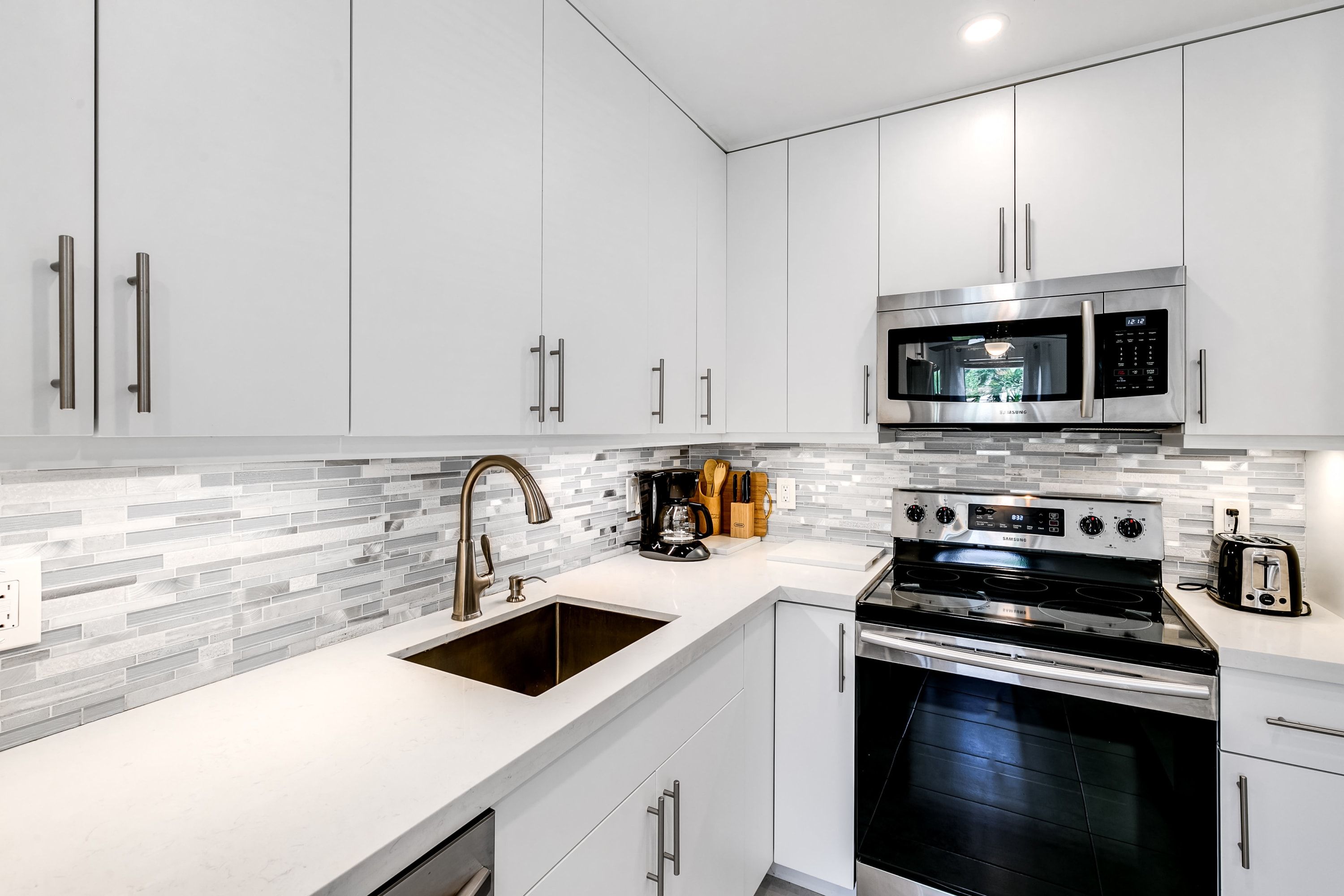 Una cocina moderna con muebles blancos, un microondas de acero inoxidable, un horno empotrado y un lavamanos con un grifo moderno.