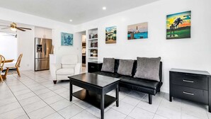 Un salon moderne avec un canapé noir, un fauteuil blanc, une table basse noire et une table d’appoint noire. Trois tableaux ornent le mur, ainsi qu’une bibliothèque et un coin cuisine avec un réfrigérateur.