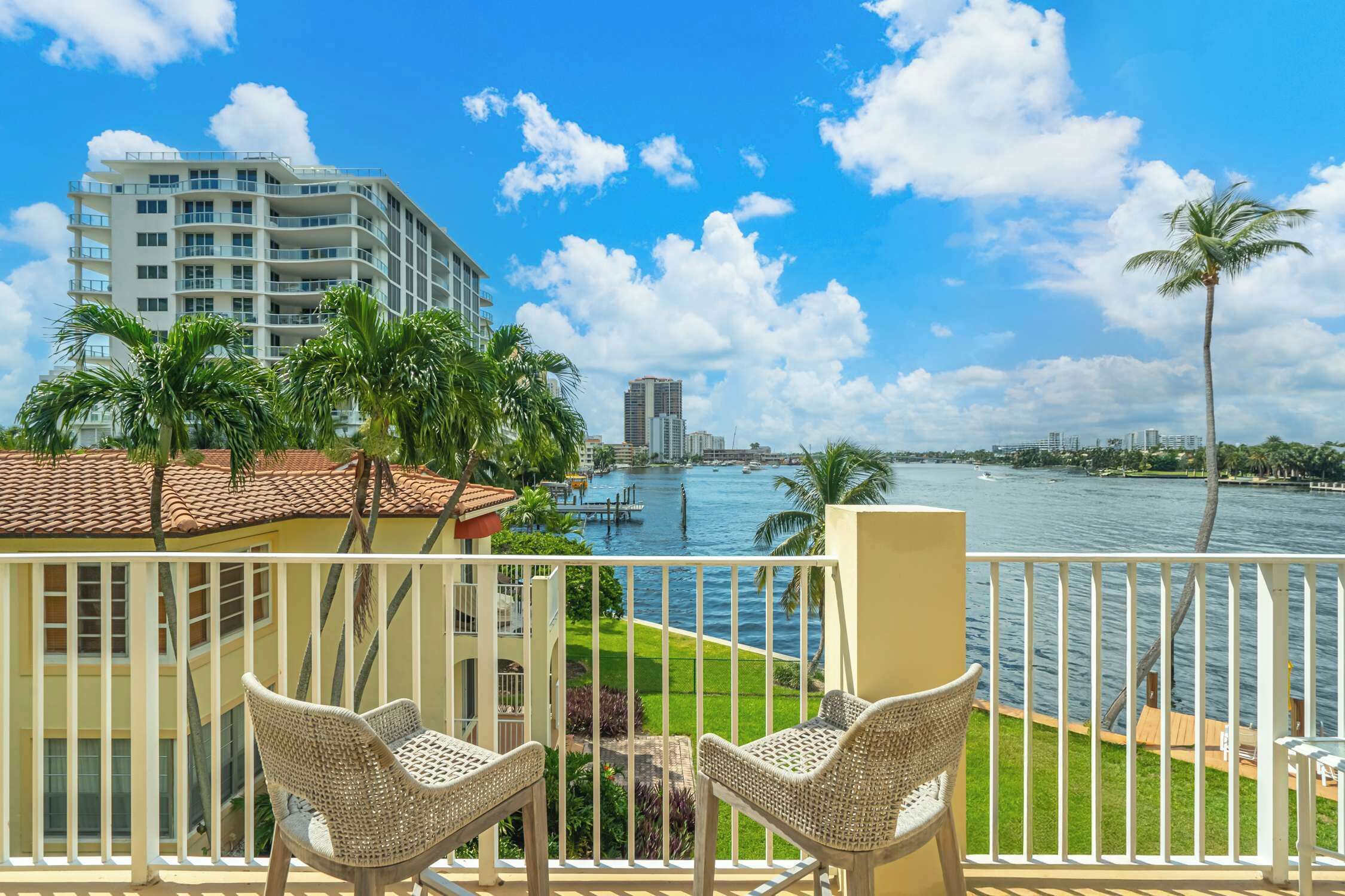 A balcony with wicker chairs overlooking a marina with boats, a city skyline, and palm trees.