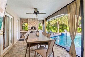Área de refeições junto à piscina, com mesa, cadeiras, ventilador de teto e vista para a piscina e vegetação tropical.