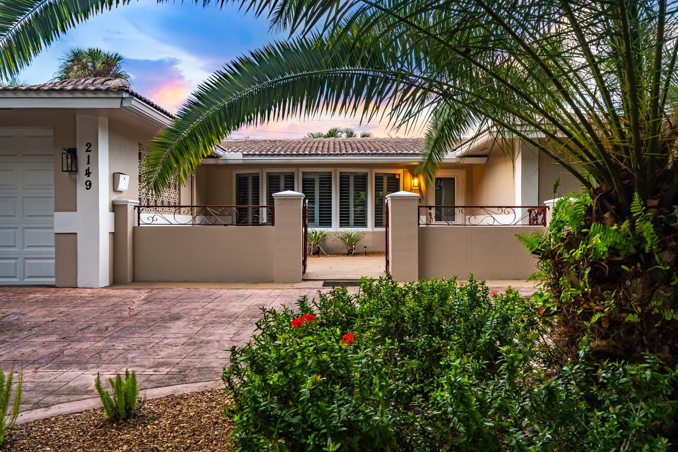 A residential property with a paved driveway, a gated entrance, and a well-maintained garden with shrubs and a palm tree.