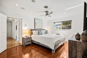 Un dormitorio con cama, mesita de noche, lámpara, ventilador de techo, ventana y vistas al exterior.