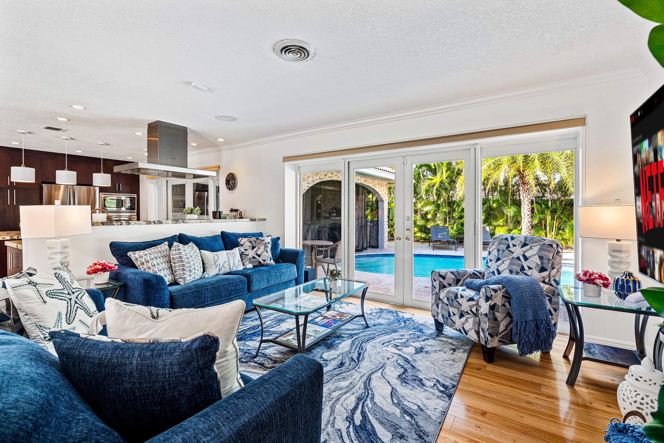 A modern living room with a blue sofa, a glass coffee table, and a poolside view.